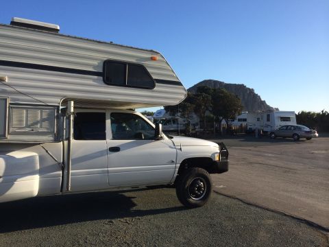 1996 Ford C-350 (Centurion Conversions) - A white pickup truck with a camper shell is parked in a lot with other RVs and a car, with a mountain in the background.