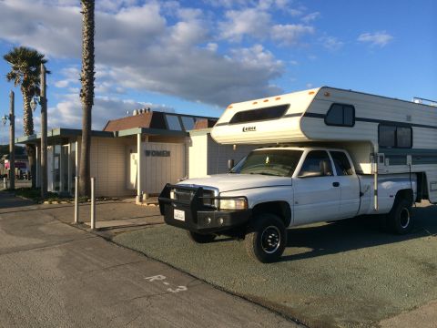 1996 Ford C-350 (Centurion Conversions) - A white pickup truck equipped with a camper shell is parked in a lot next to a building with palm trees and a blue sky overhead.