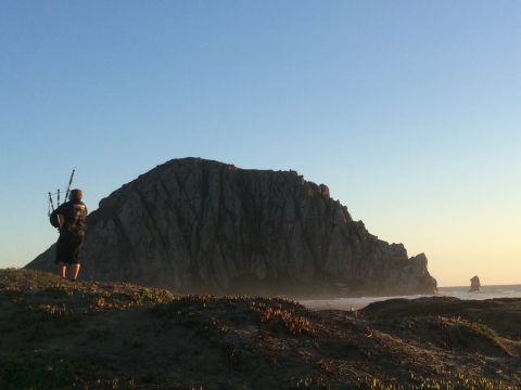 1996 Ford C-350 (Centurion Conversions) - A lone figure plays bagpipes on a coastal dune, with a prominent rock formation rising from the sea.