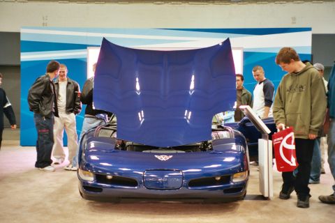 1992 Mercury Grand Marquis - A group of people gather around a blue Chevrolet Corvette C5 with its hood open at an indoor event.