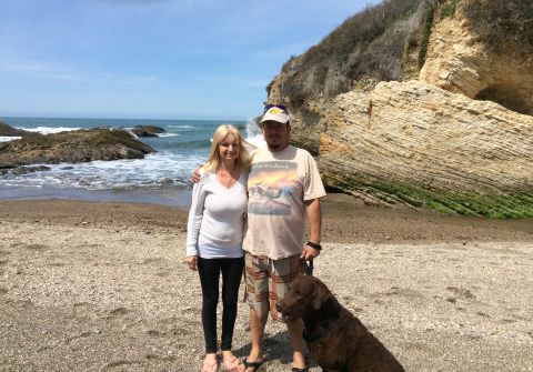 1996 Ford C-350 (Centurion Conversions) - A couple and their dog pose for a photo on a beach with waves crashing on rocks and a large cliff face.