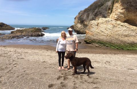 1996 Ford C-350 (Centurion Conversions) - A couple and their dog enjoy a day at the beach, with waves crashing on the shore and large rock formations in the background.