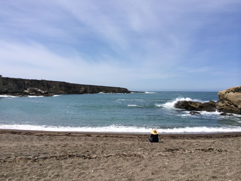 Person wearing a straw hat sits on a dark sand beach, fishing in the ocean with waves crashing on rocks