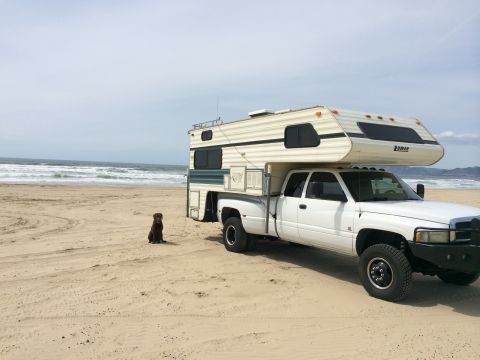 1996 Ford C-350 (Centurion Conversions) - A white dually pickup truck with a camper shell is parked on a sandy beach with waves in the background. A dog sits nearby.