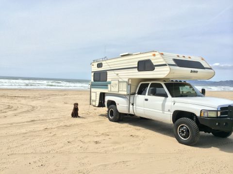1996 Ford C-350 (Centurion Conversions) - A white pickup truck with a camper is parked on a sandy beach with the ocean in the background, while a dog sits patiently nearby.