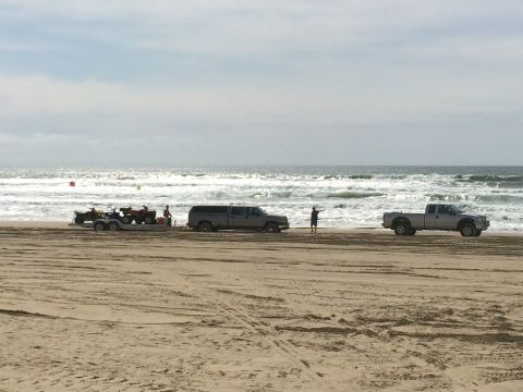 1996 Ford C-350 (Centurion Conversions) - Two pickup trucks and a trailer with ATVs are parked on a beach with waves in the background.