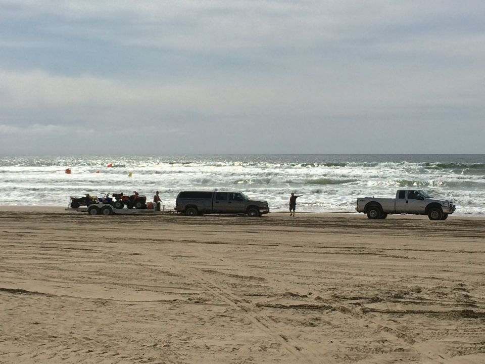 Silver pickup truck towing ATVs on a trailer, with another pickup truck nearby on a beach