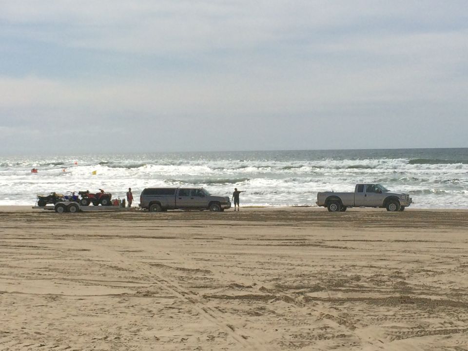 Two pickup trucks, one towing ATVs on a trailer, parked on a sandy beach near the ocean