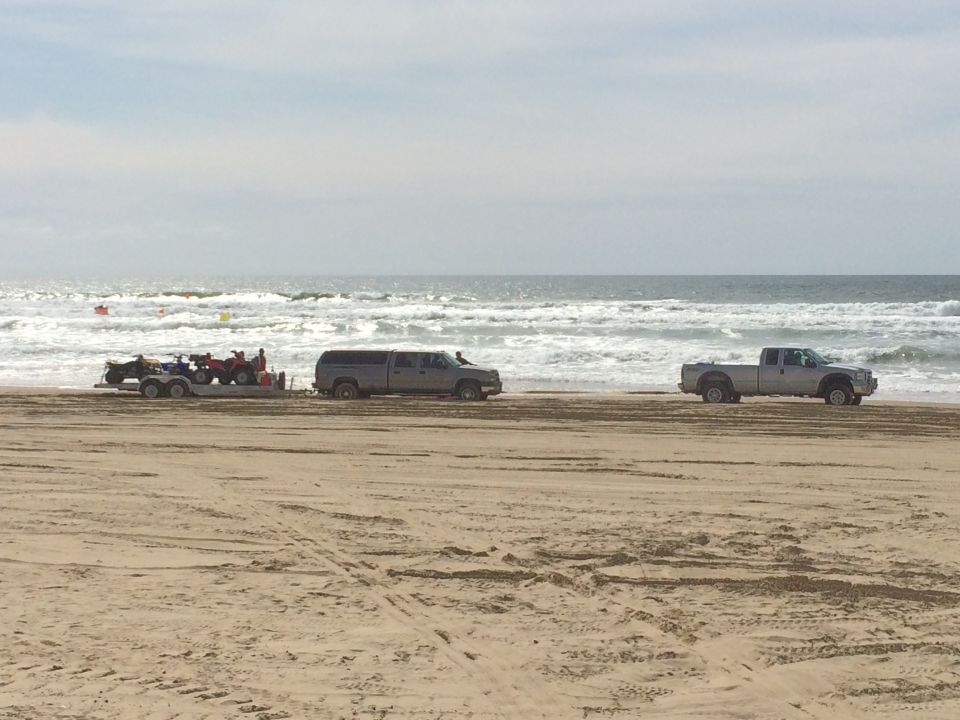 Two pickup trucks and a trailer with ATVs on a sandy beach with the ocean in the background