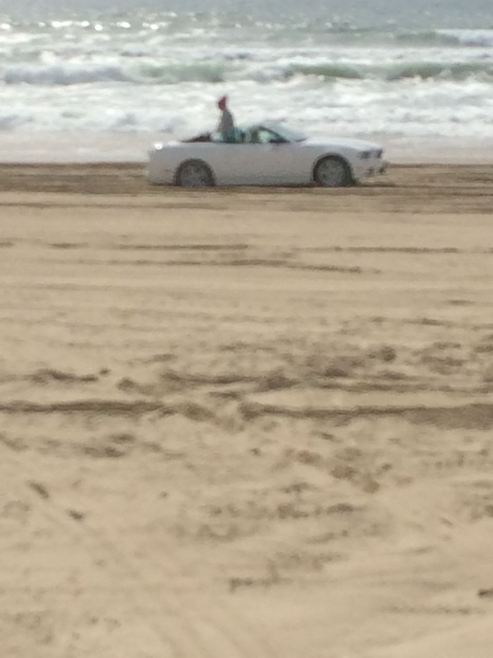 White convertible with a person inside driving on a sandy beach near the ocean