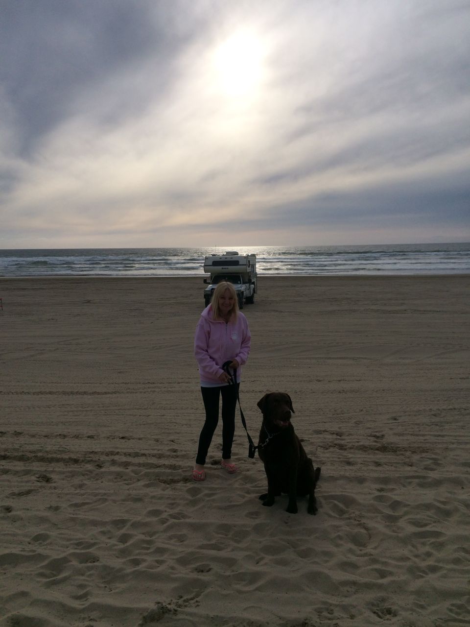 Woman holding a leash with a dog sitting on the sand, RVs parked on the beach near the ocean