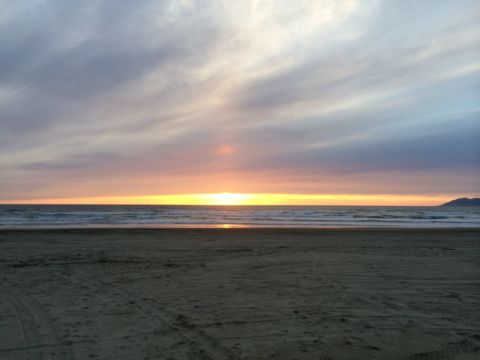 1996 Ford C-350 (Centurion Conversions) - sun sets low over the ocean horizon from a sandy beach marked with tire tracks under a streaked cloudy sky.