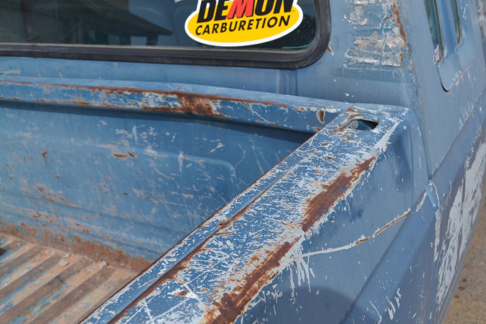 Blue pickup truck bed with rust and scratches. Sticker on rear window says 'DEMON CARBURETION'