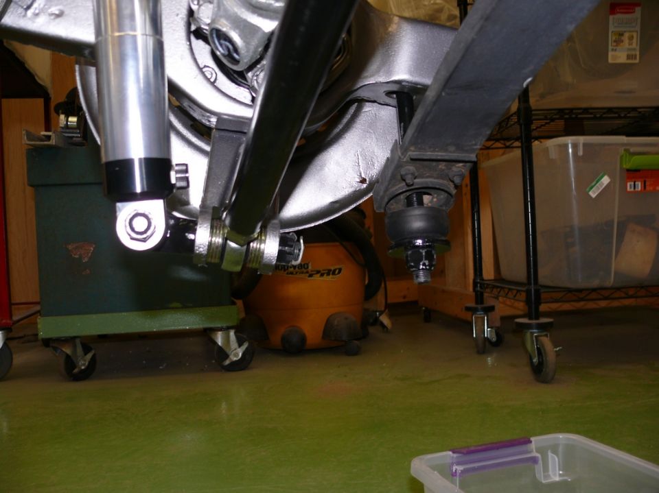 Close-up of car suspension components, including a shock absorber and control arm, with a shop vacuum and storage shelves in the background