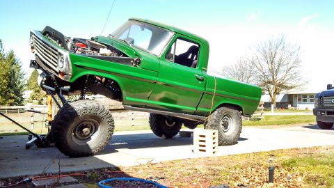 1967 Ford F-250 - A modified green pickup truck is undergoing maintenance outdoors, with its front end elevated by a jack.