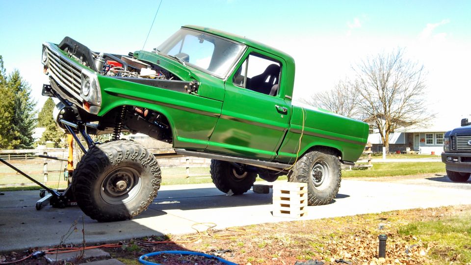 Green lifted pickup truck with hood open, jacked up on front end. Another dark pickup truck is parked nearby