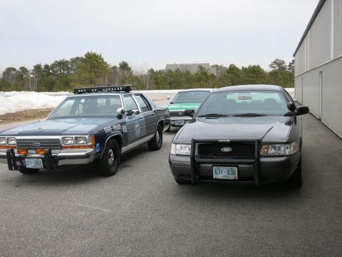 1991 Ford LTD Crown Victoria - A dark grey Ford Crown Victoria and a blue Ford police car are parked in front of a building.