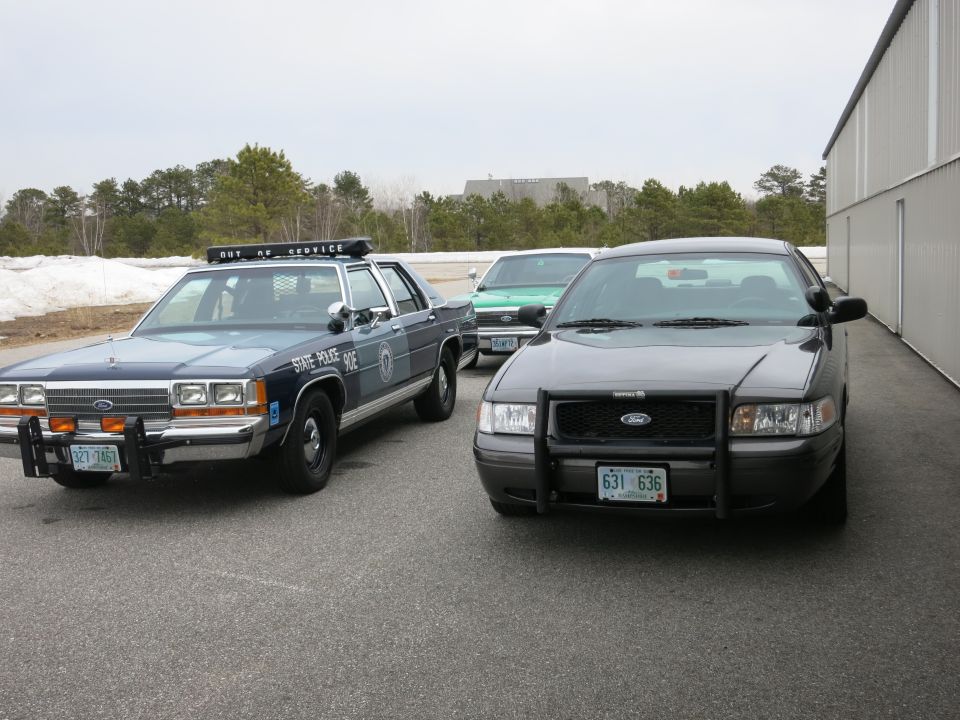Two police cars, a dark grey Ford Crown Victoria and a blue Ford, parked outside a building