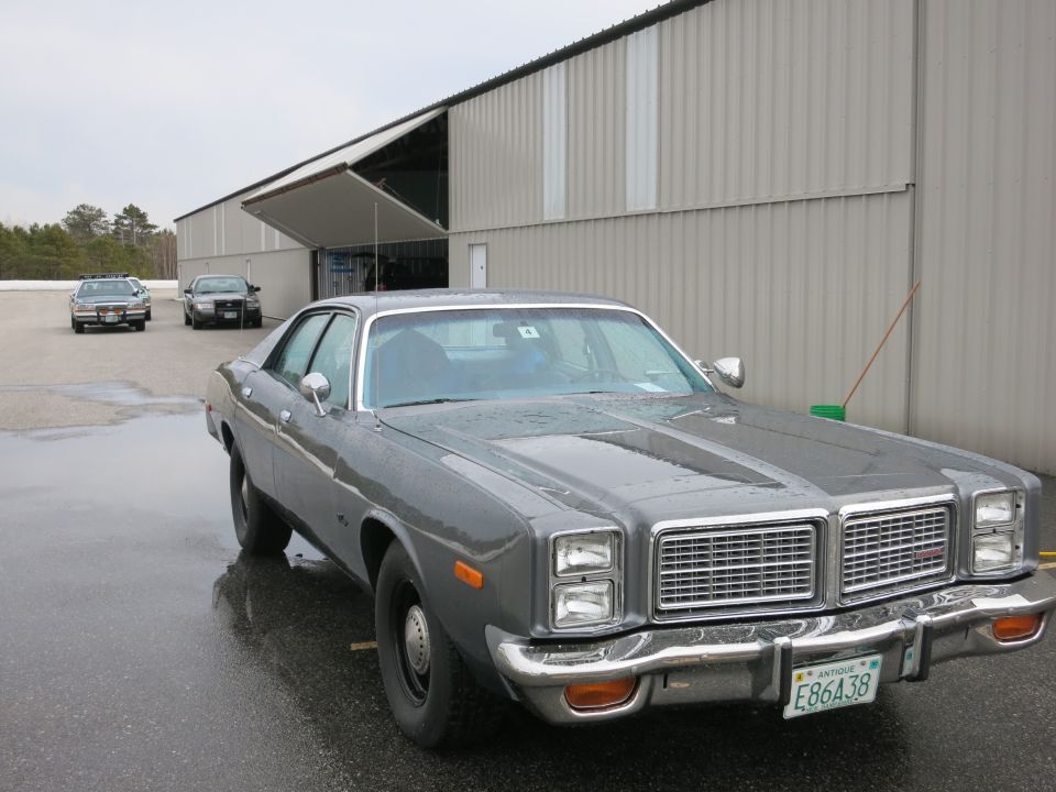 1977 Dodge Monaco parked in front of a hangar, two police cars in background