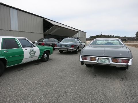 1991 Ford LTD Crown Victoria - Several vehicles, including police cars, are parked outside a large industrial building.