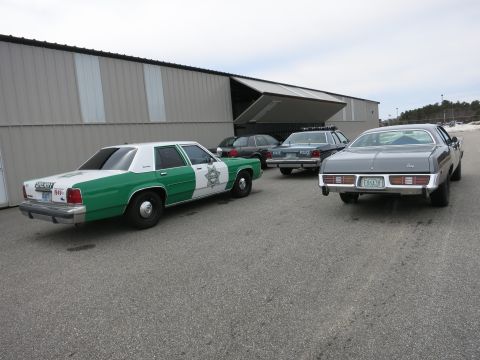 1991 Ford LTD Crown Victoria - A fleet of older vehicles, including a sheriff's car and a sedan, are parked outside a large industrial building.