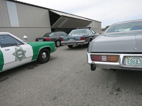 1991 Ford LTD Crown Victoria - Multiple police vehicles and a grey car are parked outside a hangar.