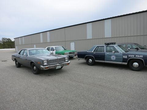 1991 Ford LTD Crown Victoria - Three sedans, including a gray 1977 model and a state police car, are parked outside a large building.