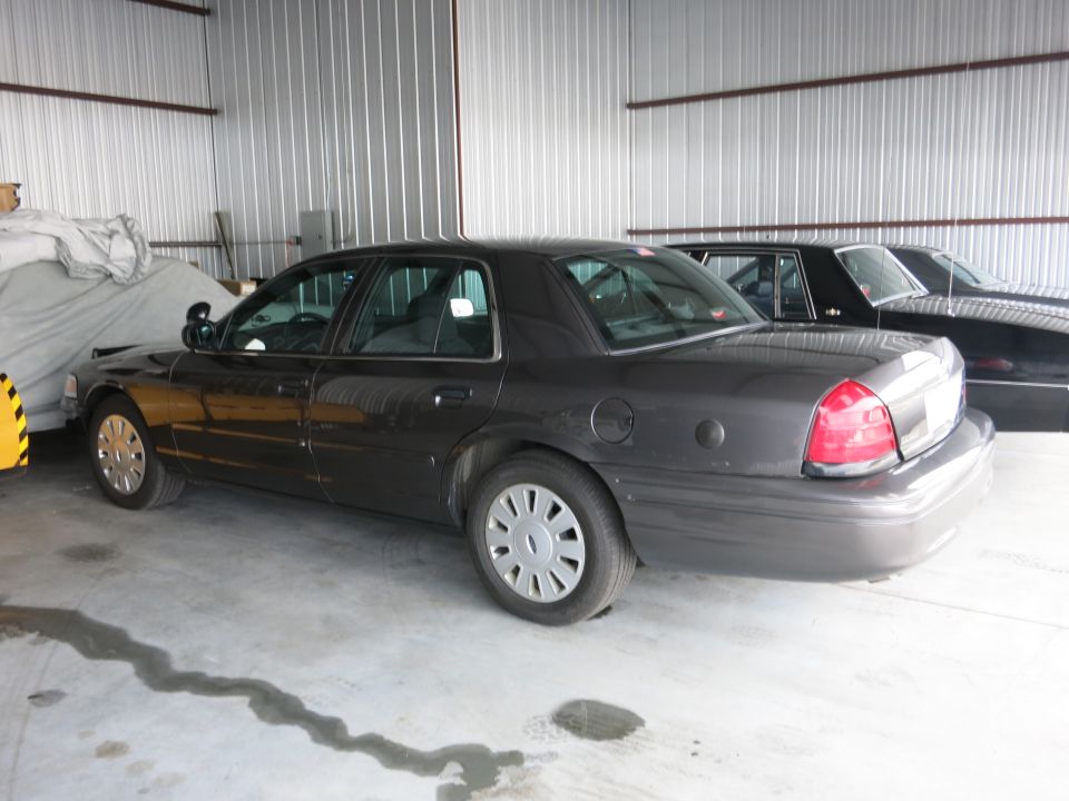 Dark gray Ford Crown Victoria parked in a garage next to another car. It has a police-style side mirror