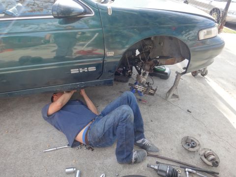 1993 Ford Taurus - A mechanic works on the front suspension of a green 1993 Ford Taurus SHO, with parts laid out on the ground.
