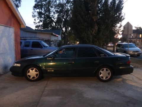 1993 Ford Taurus - A dark green sedan is parked in a driveway next to a garage, with a blue pickup truck behind it.