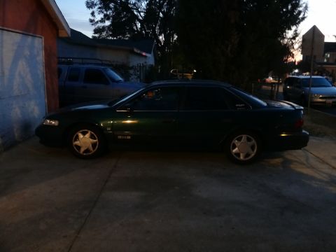 1993 Ford Taurus - A dark green sedan is parked on a concrete driveway at dusk, with a blue pickup truck visible behind it.