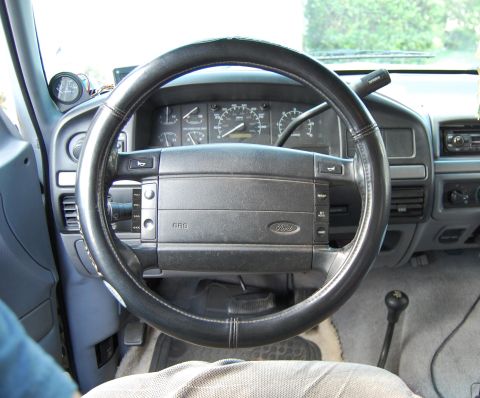 1994 Ford Bronco - Driver's-seat view of a blue Ford truck interior showing the four-spoke SRS airbag wheel with a laced-on leather cover, cruise and audio sw…