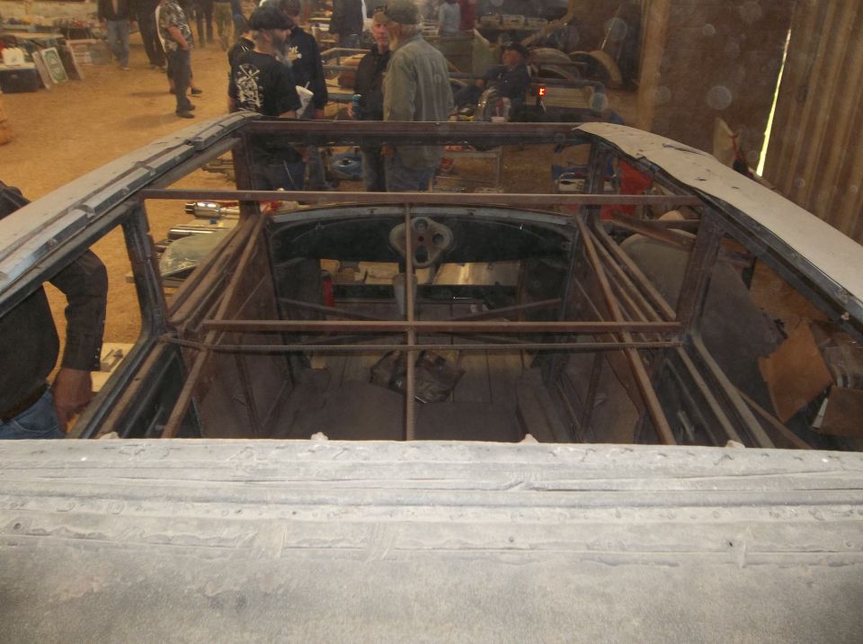 Interior of a car under construction, showing a roll cage and bare metal frame. People are visible in the background