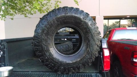 1985 Jeep CJ7 - A large spare tire is positioned in the bed of a black pickup truck, with a red pickup truck visible to the right.