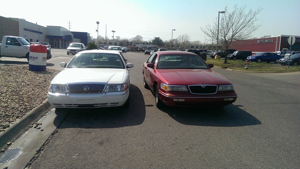 Two Mercury Grand Marquis sedans parked side-by-side. The one on the left is white, and the one on the right is red. Both are facing forward