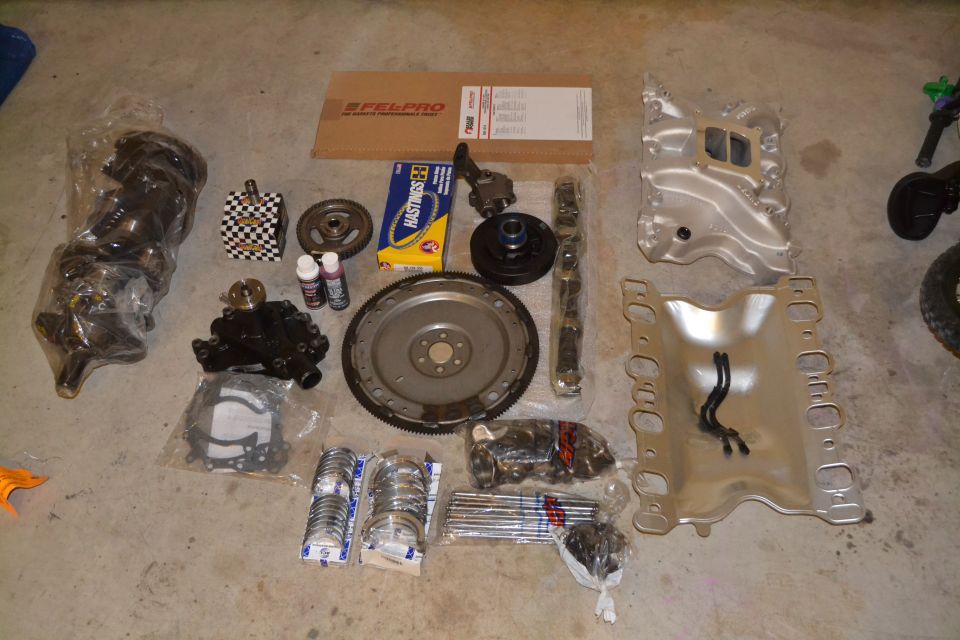 Engine parts including crankshaft, intake manifold, exhaust manifolds, pistons, bearings, and gaskets laid out on concrete floor