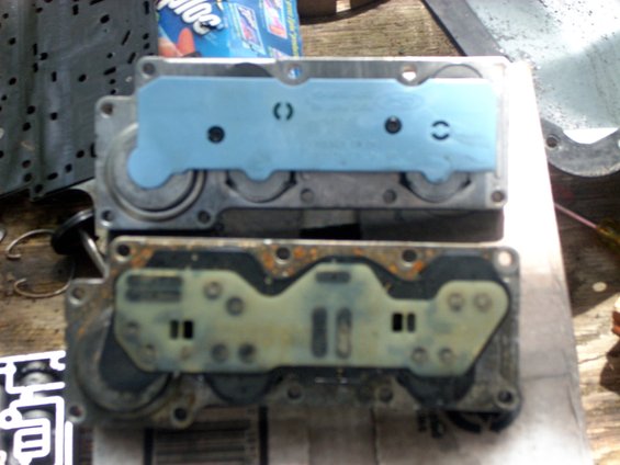 Two transmission valve bodies on a workbench, one with blue gasket plate, one dirty with rust