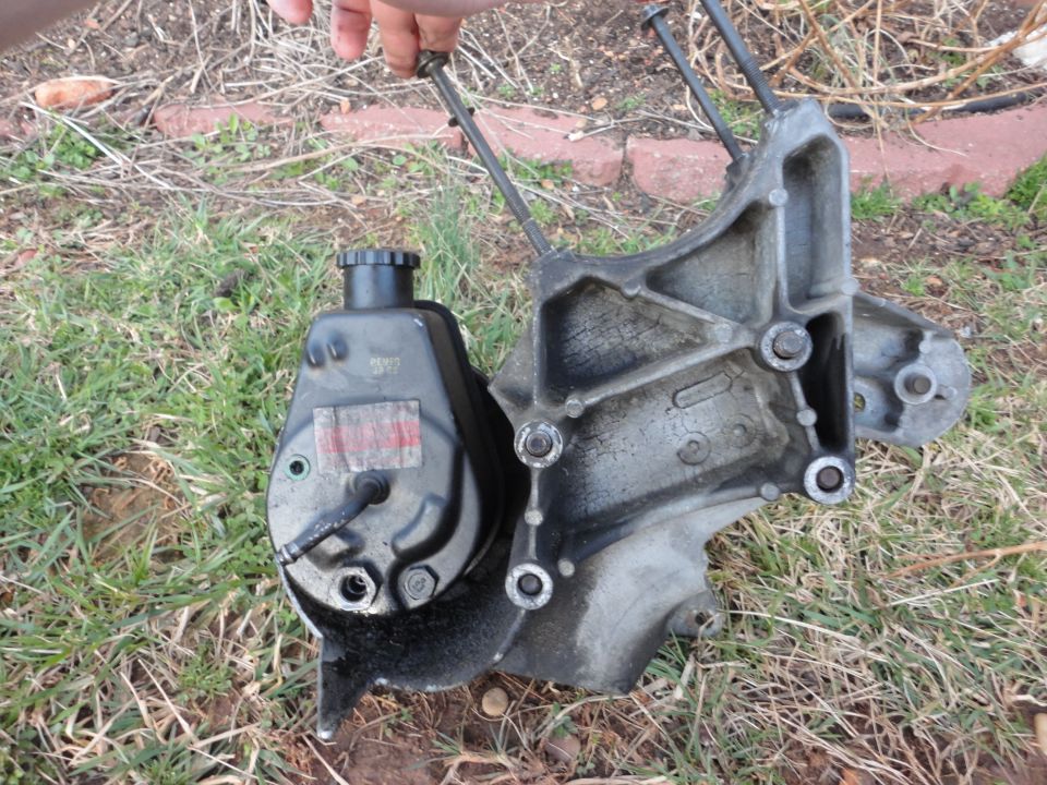 Dirty power steering pump and reservoir attached to aluminum bracket with long bolts, held over grass