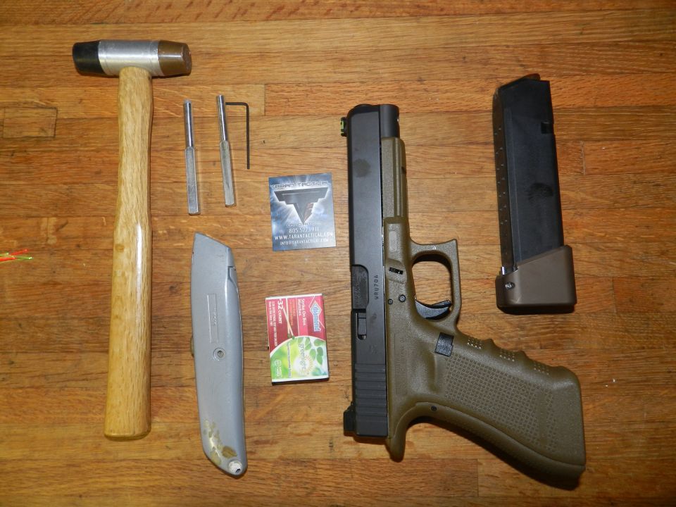 Handgun with extended magazine, utility knife, hammer, punches, and a small box on a wooden surface