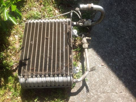 1976 Ford Ranchero - A car's air conditioning evaporator core, a metal component with fins and attached hoses, rests outdoors on grass and pavement.