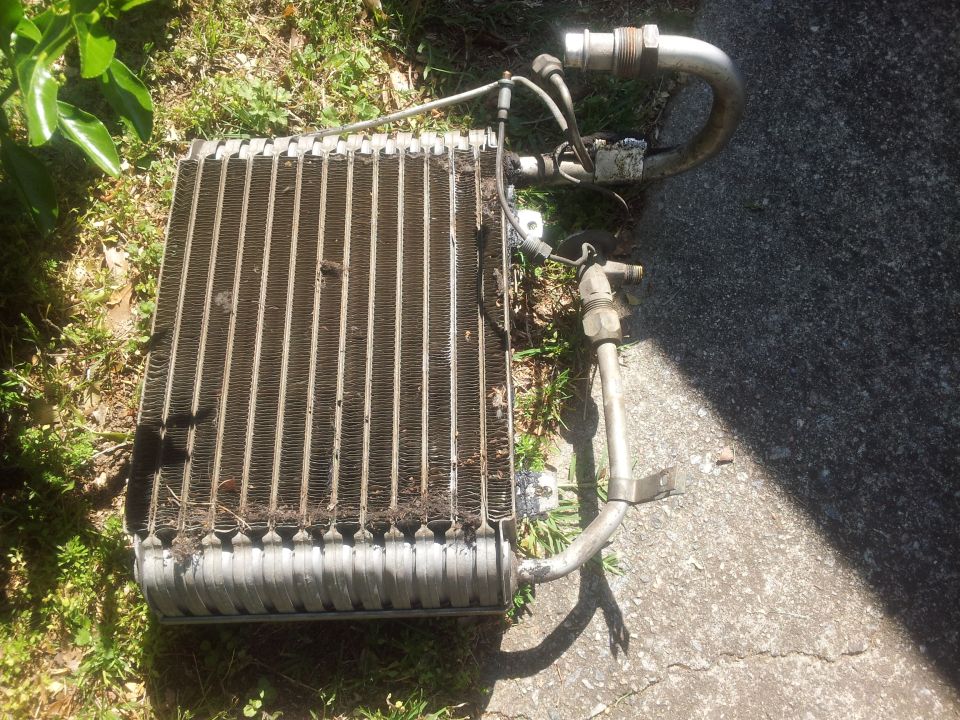 Car AC evaporator core with metal fins and hoses, lying on grass and asphalt