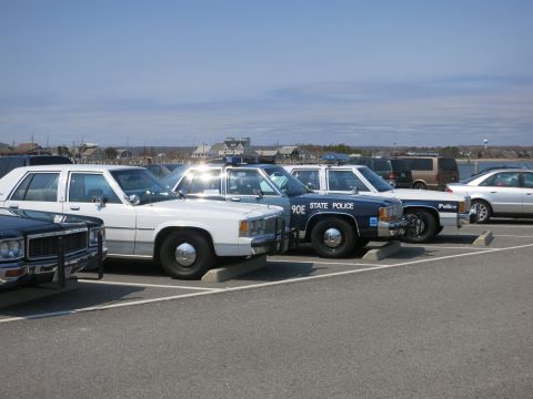 1991 Ford LTD Crown Victoria - row of vintage police cars parked in a waterfront lot, including a blue 90E STATE POLICE Ford cruiser and white sedans.