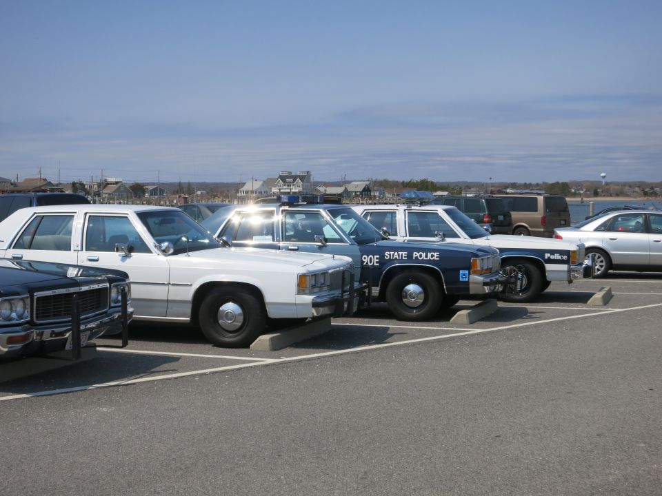 Row of vintage police cars in a waterfront lot, blue STATE POLICE cruiser
