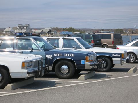 1991 Ford LTD Crown Victoria - Close view of parked police cruisers in a lot, centered on a blue 90E STATE POLICE Ford with a lightbar and push bumper.