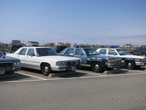 1991 Ford LTD Crown Victoria - row of parked vintage police cars in a lot, including a blue 90E STATE POLICE Ford cruiser flanked by white sedans.
