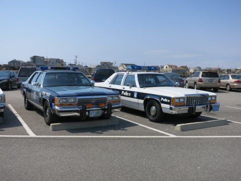 1991 Ford LTD Crown Victoria - Two vintage Ford police cruisers parked in a lot, a blue cruiser with push bumper beside a black-and-white car marked Police.