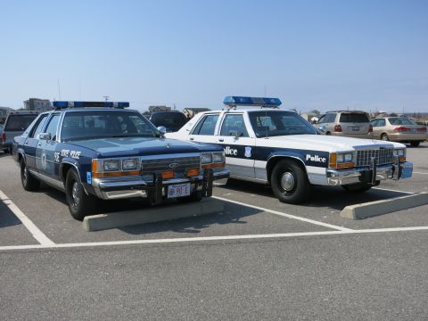 1991 Ford LTD Crown Victoria - blue 90E STATE POLICE Ford cruiser parked beside a black-and-white Ford marked Police in a sunny lot.