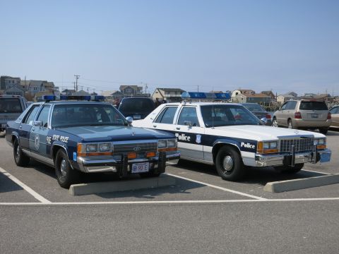 1991 Ford LTD Crown Victoria - blue 90E STATE POLICE Ford cruiser with push bumper parked beside a black-and-white Ford marked Police near coastal houses.