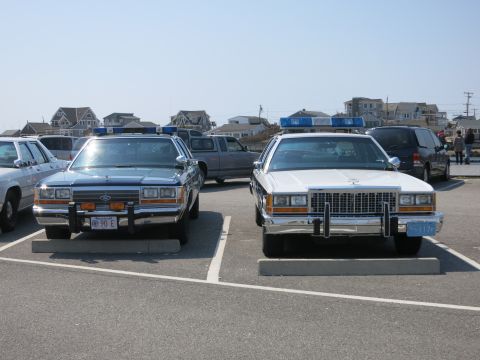 1991 Ford LTD Crown Victoria - Front view of two parked Ford police cruisers, a blue 90E cruiser with Massachusetts plate beside a white-over-blue police car.