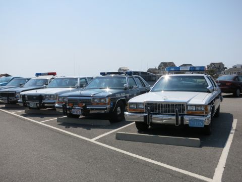 1991 Ford LTD Crown Victoria - lineup of classic police cruisers, including a State Police sedan, parked diagonally in a coastal parking lot.
